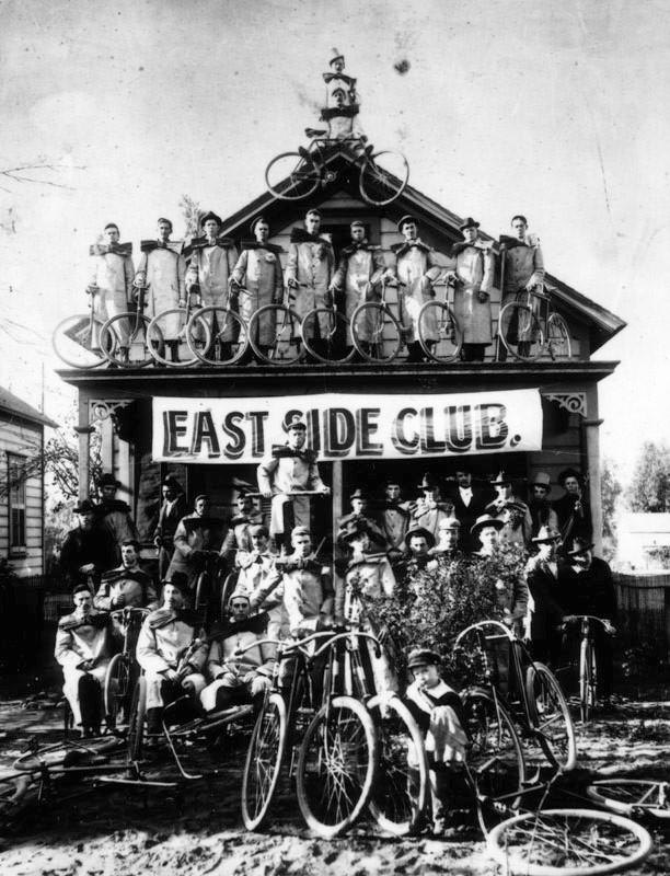 Group photo of the East Side Cycle Club, showing a banner for the club, taken on the beach at Santa Monica near the old North Beach Bath House. The riders are wearing overcoats an over-sized bows.