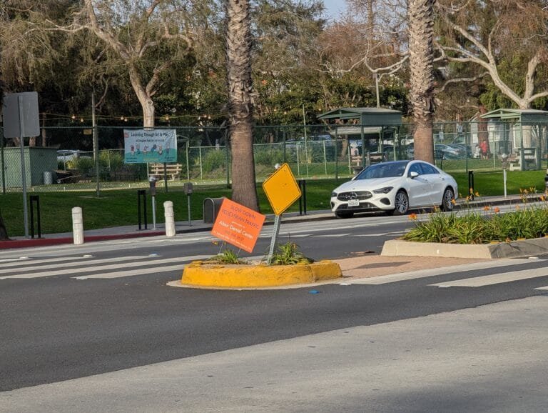 A crosswalk at Wilshire and Chelsea with Douglas Park in the background. Someone has placed a pedestrian safety sign that also advertises a local dental clinic.