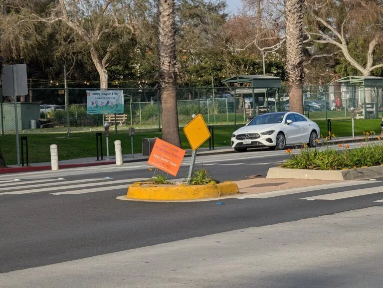 A crosswalk at Wilshire and Chelsea with Douglas Park in the background. Someone has placed a pedestrian safety sign that also advertises a local dental clinic.